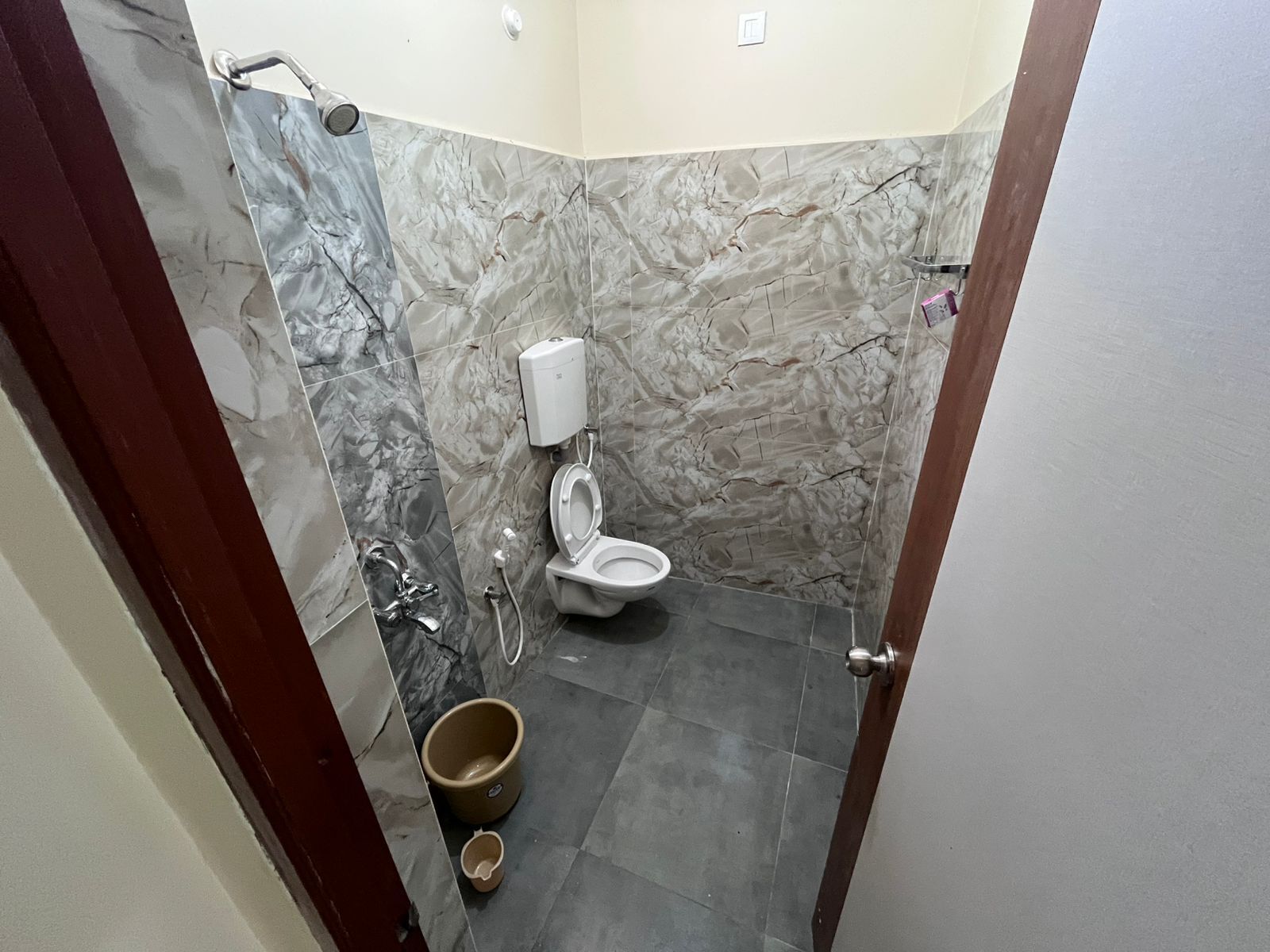 A bathroom with a toilet, marble walls, a shower, and a bucket on the floor.