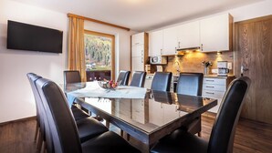 Een moderne eetruimte met een glazen tafel, zwarte leren stoelen, een flatscreen televisie, en een keuken met houten kasten en uitzicht op een bos.
