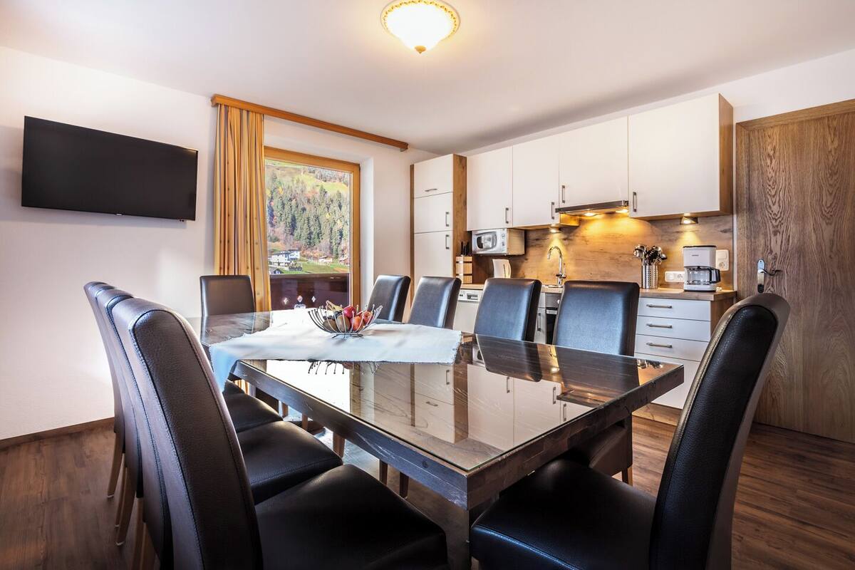 A modern dining area with a glass table, black leather chairs, a flat-screen TV, and a kitchen with wooden cabinets and a view of a forest.