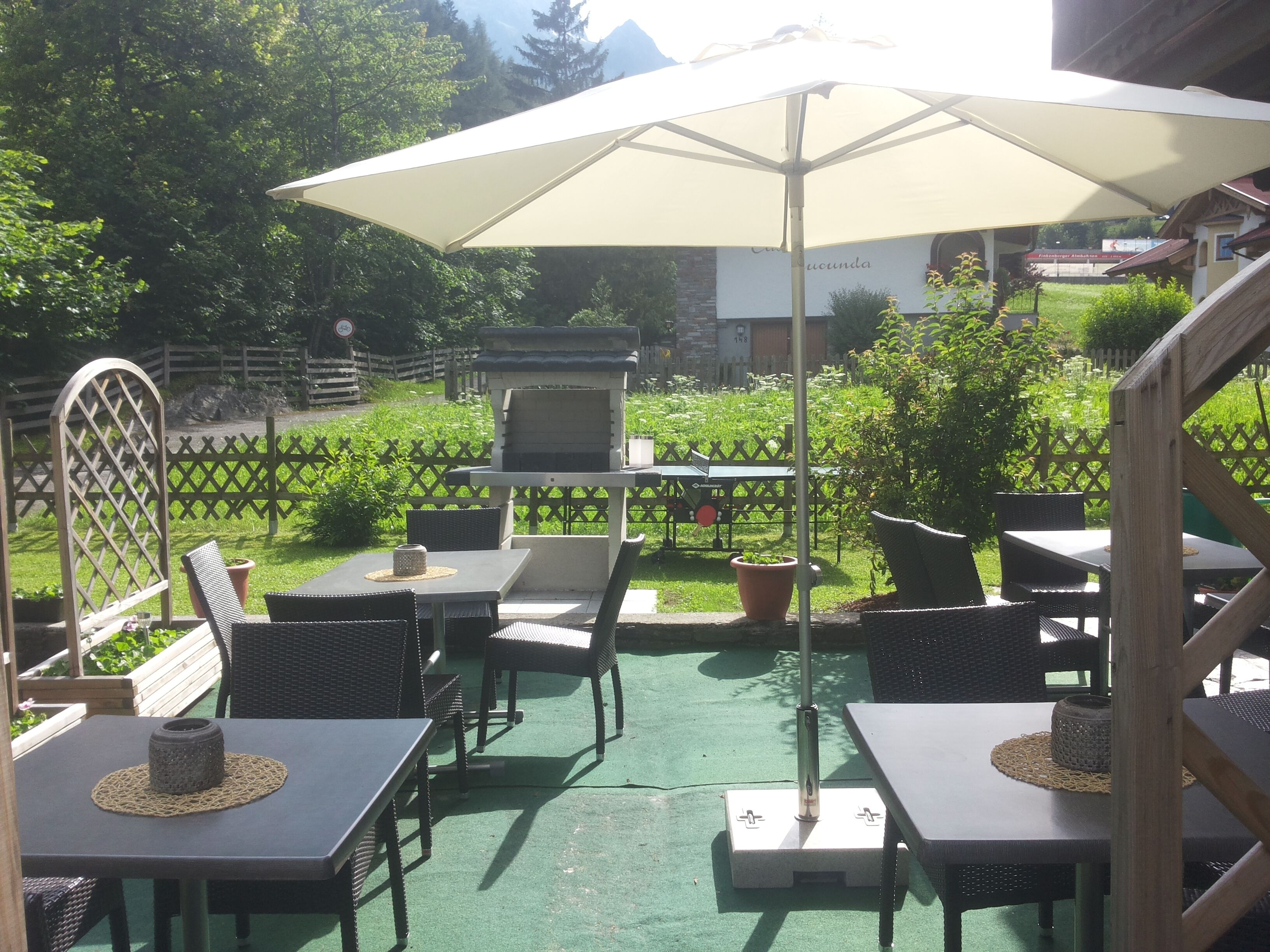 An outdoor patio area with tables and chairs, a barbecue grill, and a large umbrella providing shade.