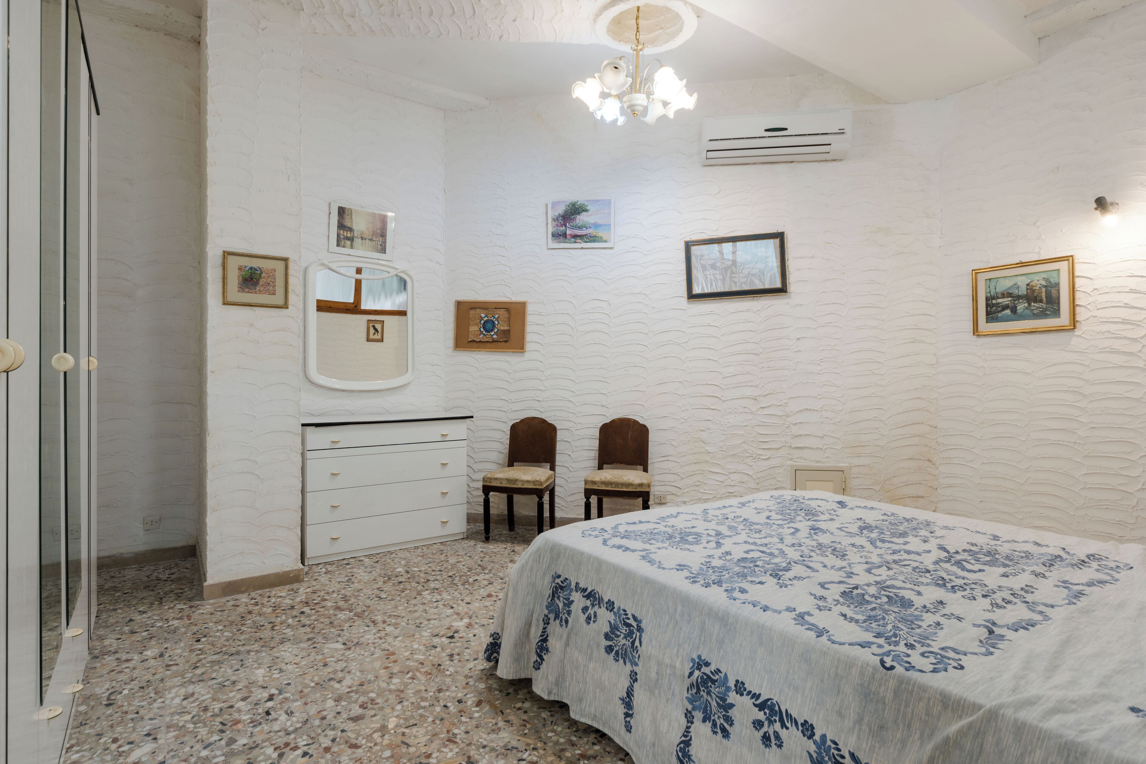 A bedroom with a bed, two chairs, a dresser, and wall-mounted air conditioning unit.