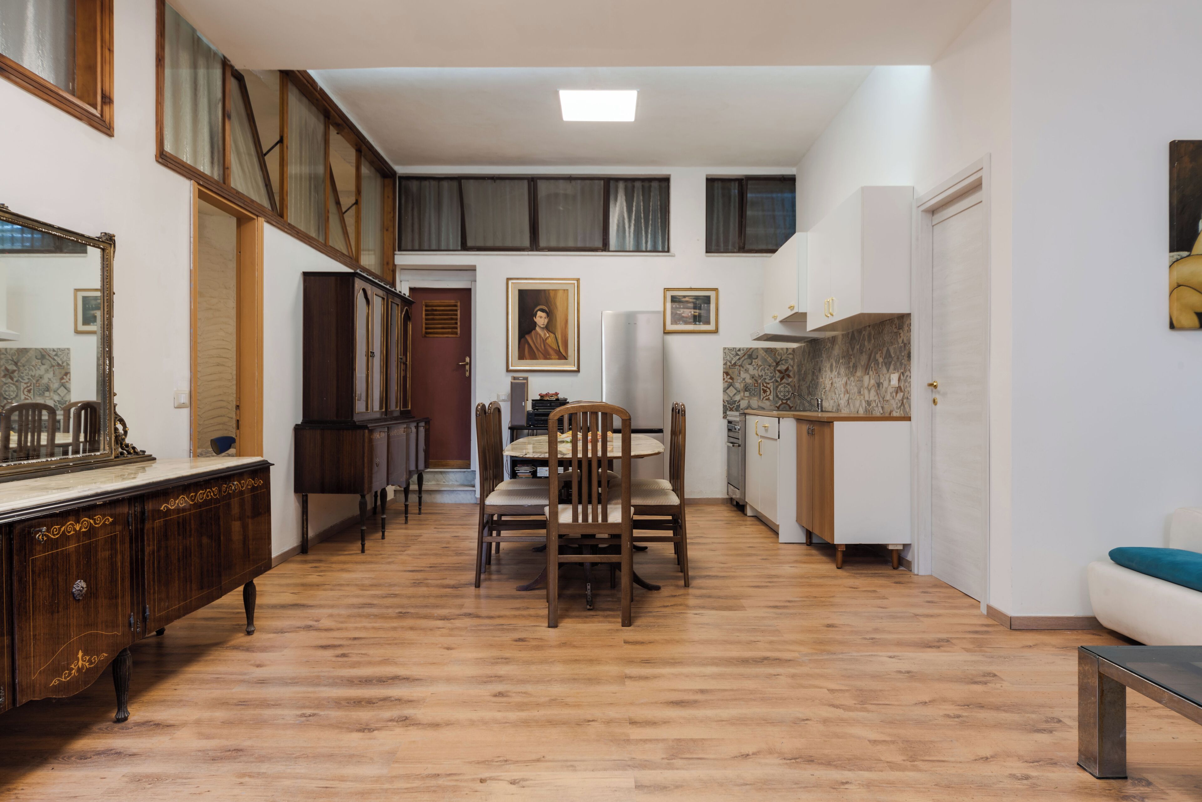 A spacious living area with a wooden floor, a dining table with chairs, a kitchen area with cabinets, and a large mirror on the left.