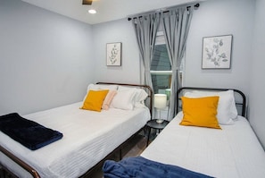Une chambre d’hôtel avec deux lits simples, chacun doté d’un matelas blanc et d’une couverture bleue. Deux œuvres d’art encadrées ornent les murs, il y a une fenêtre avec des rideaux et une petite table avec une lampe placée entre les lits.