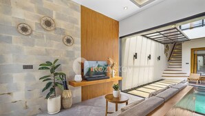A modern living room with a stone wall, wooden paneling, a flat-screen TV, a small table, a sofa, and a pool area. - Oslen Villa by Kozystay - Canggu (Canggu)