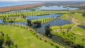 Vue aérienne d’un terrain de golf avec plusieurs étangs, des palmiers et une silhouette de ville en arrière-plan.