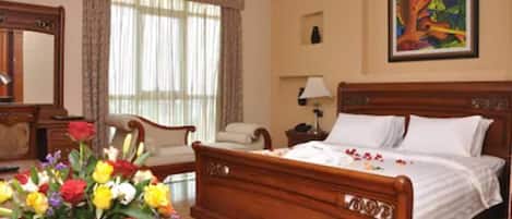A bedroom with a wooden bed, a nightstand, a chair, a desk, a desk lamp, a window with curtains, and a painting on the wall.