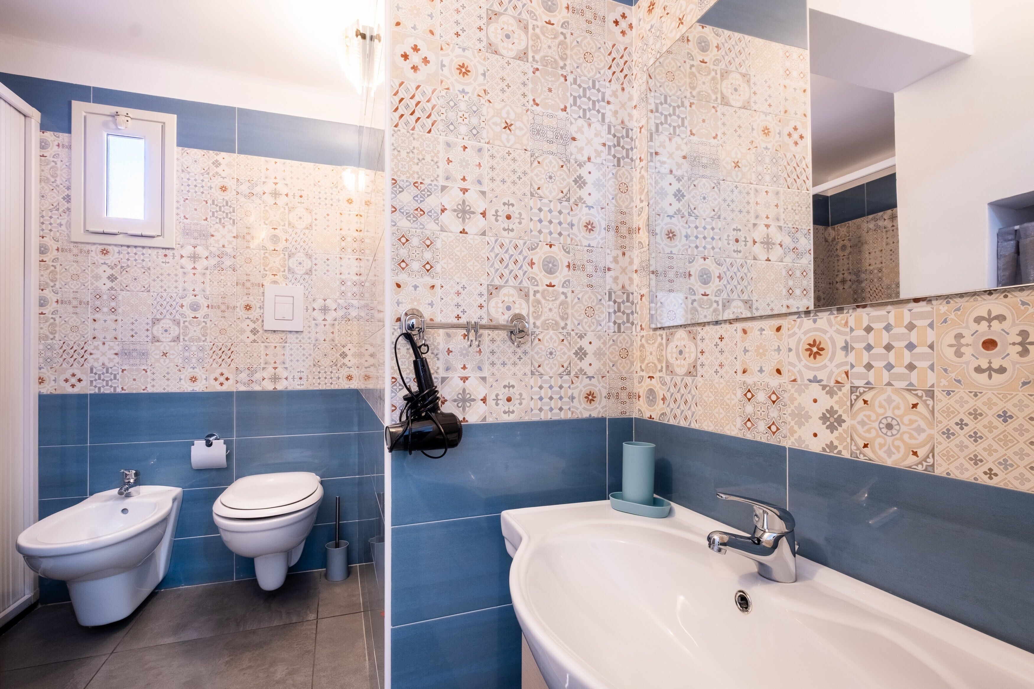 A bathroom with a bidet, toilet, and sink, featuring patterned tiles and a mirror.