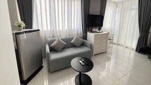 Une chambre d’hôtel moderne avec un canapé gris, une petite table ronde avec une télécommande, un minibar, un téléviseur à écran plat et un bureau avec une chaise.