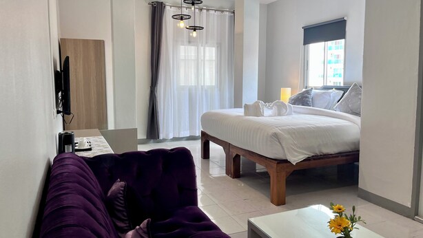 Une chambre d’hôtel avec un lit, un bureau équipé d’un ordinateur, un canapé violet et une petite table agrémentée de fleurs.