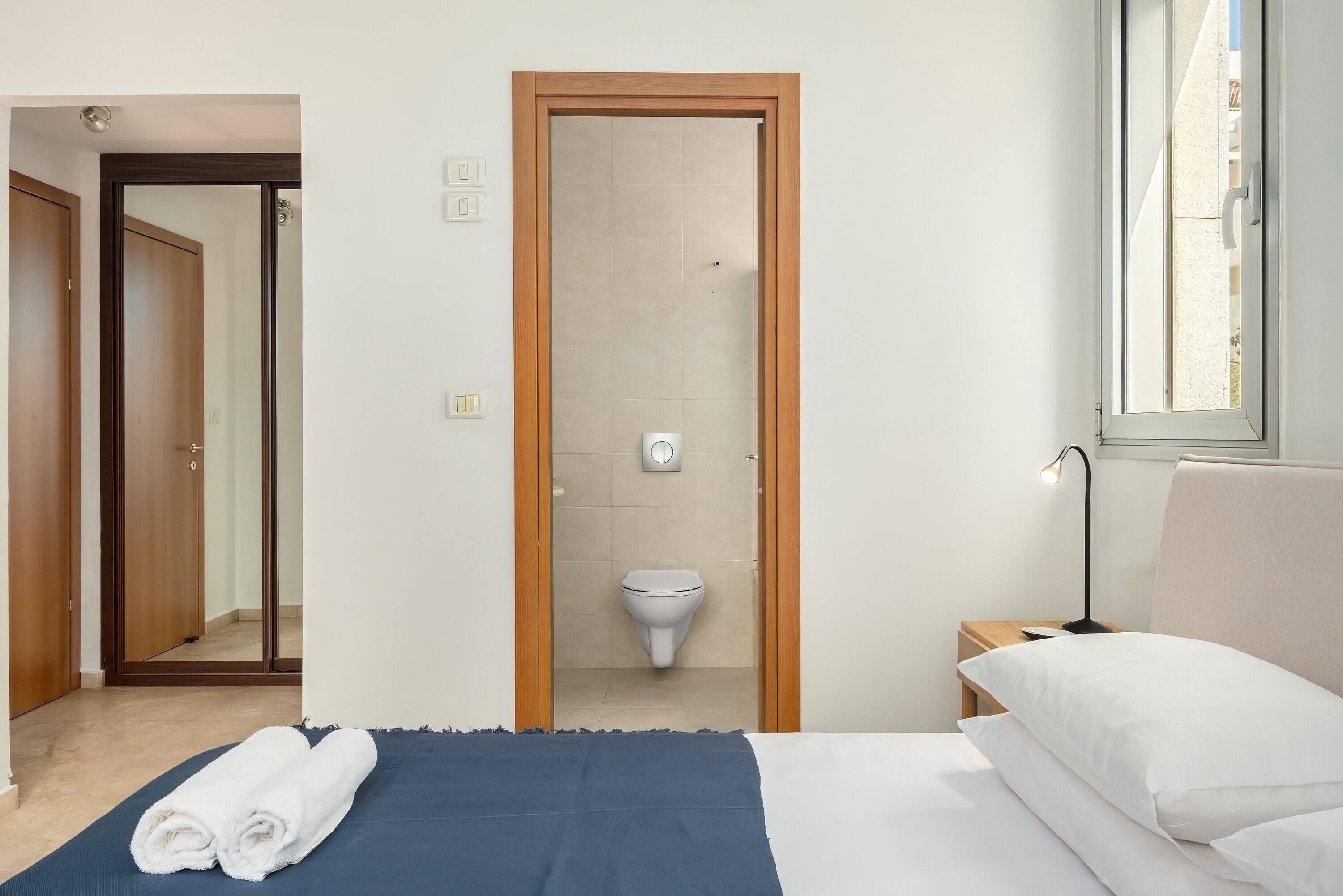 A bedroom with a bed, a nightstand, a bedside lamp, a window, and a view of the bathroom.
