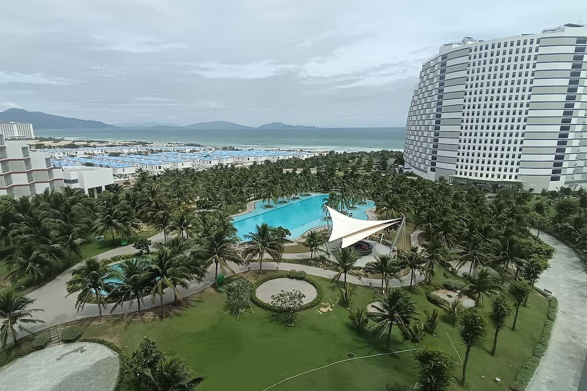 Ett semesterhotell med en central pool omgiven av palmer, en stor hotellbyggnad och utsikt över havet.