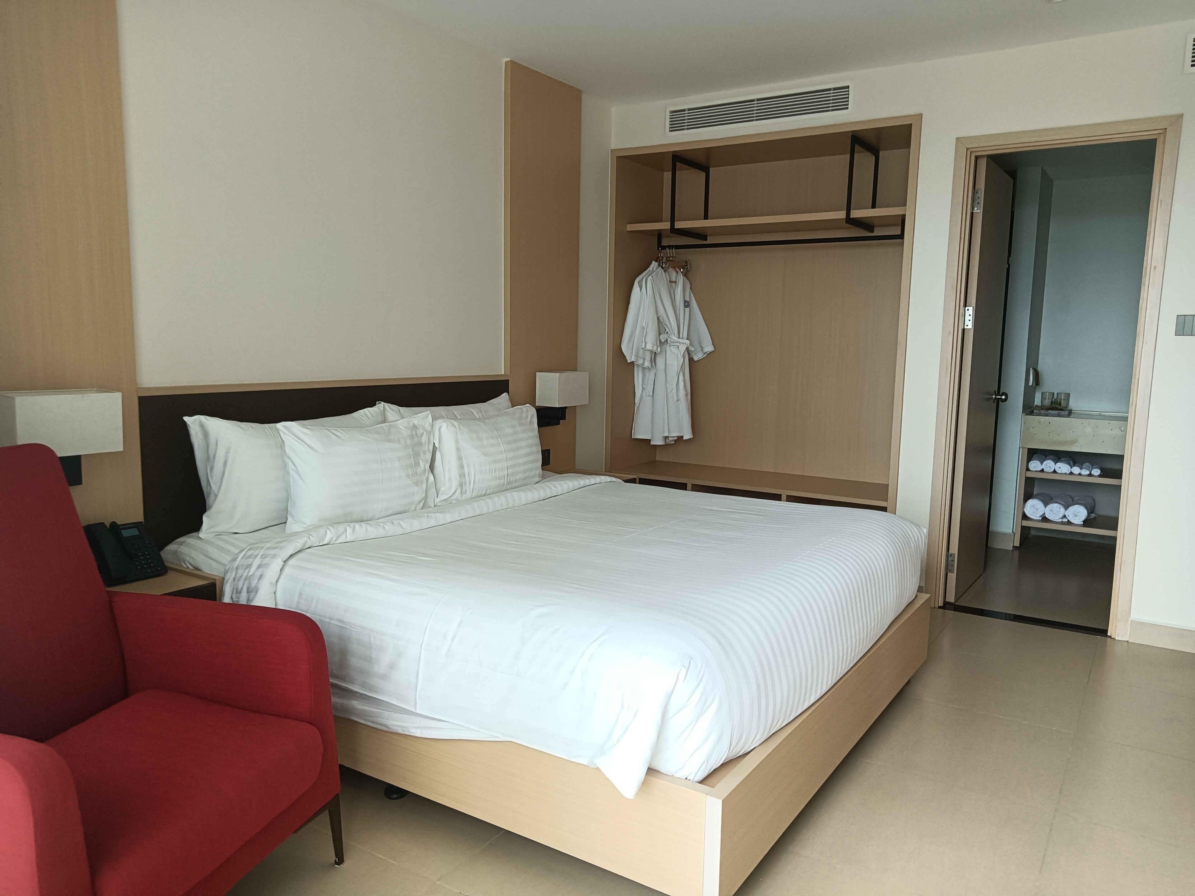 Une chambre d’hôtel avec un lit, une chaise rouge, un peignoir suspendu à un porte-manteau et une salle de bain avec des étagères.