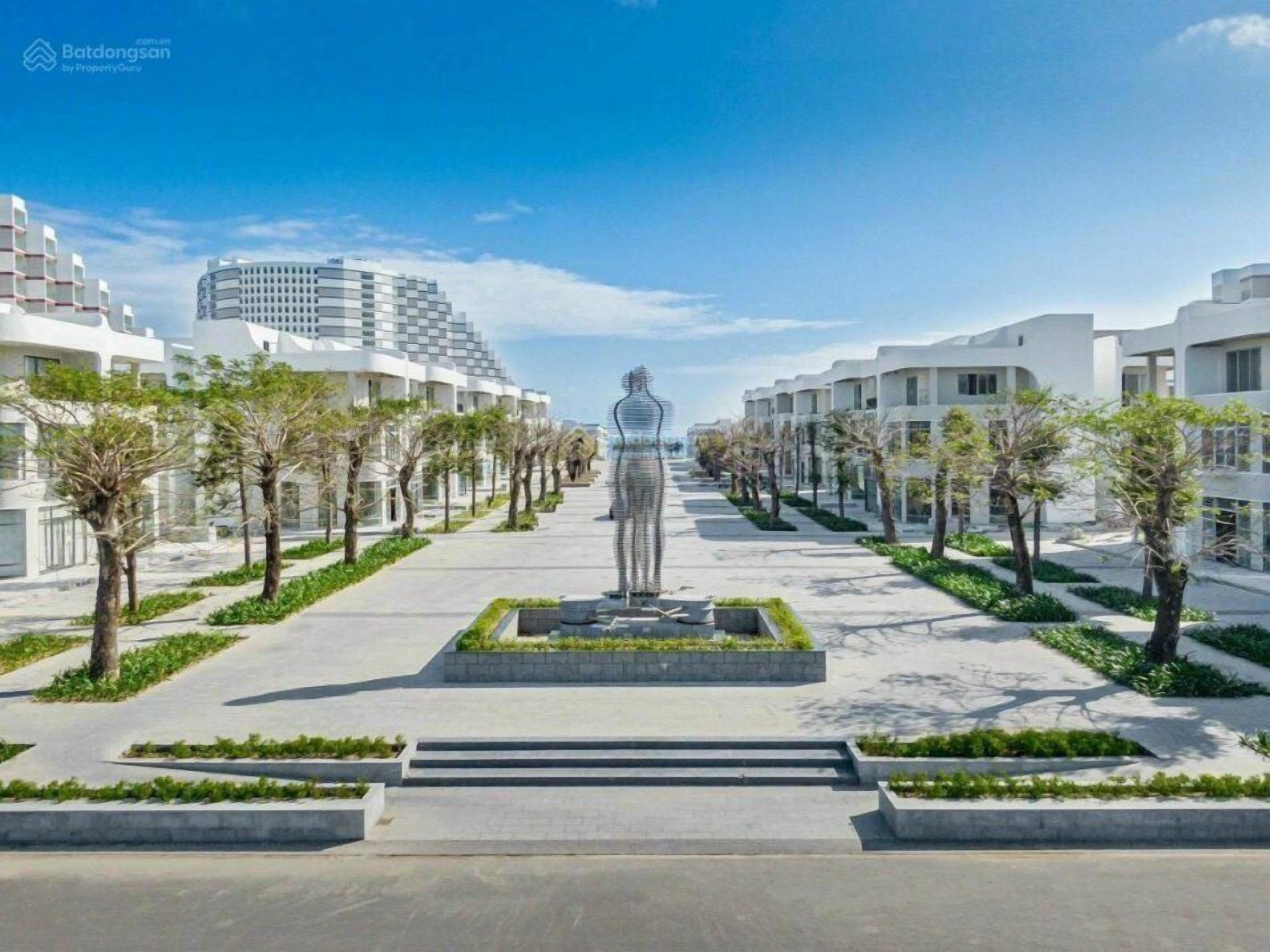 A modern urban street with a central statue, lined with trees and white buildings.