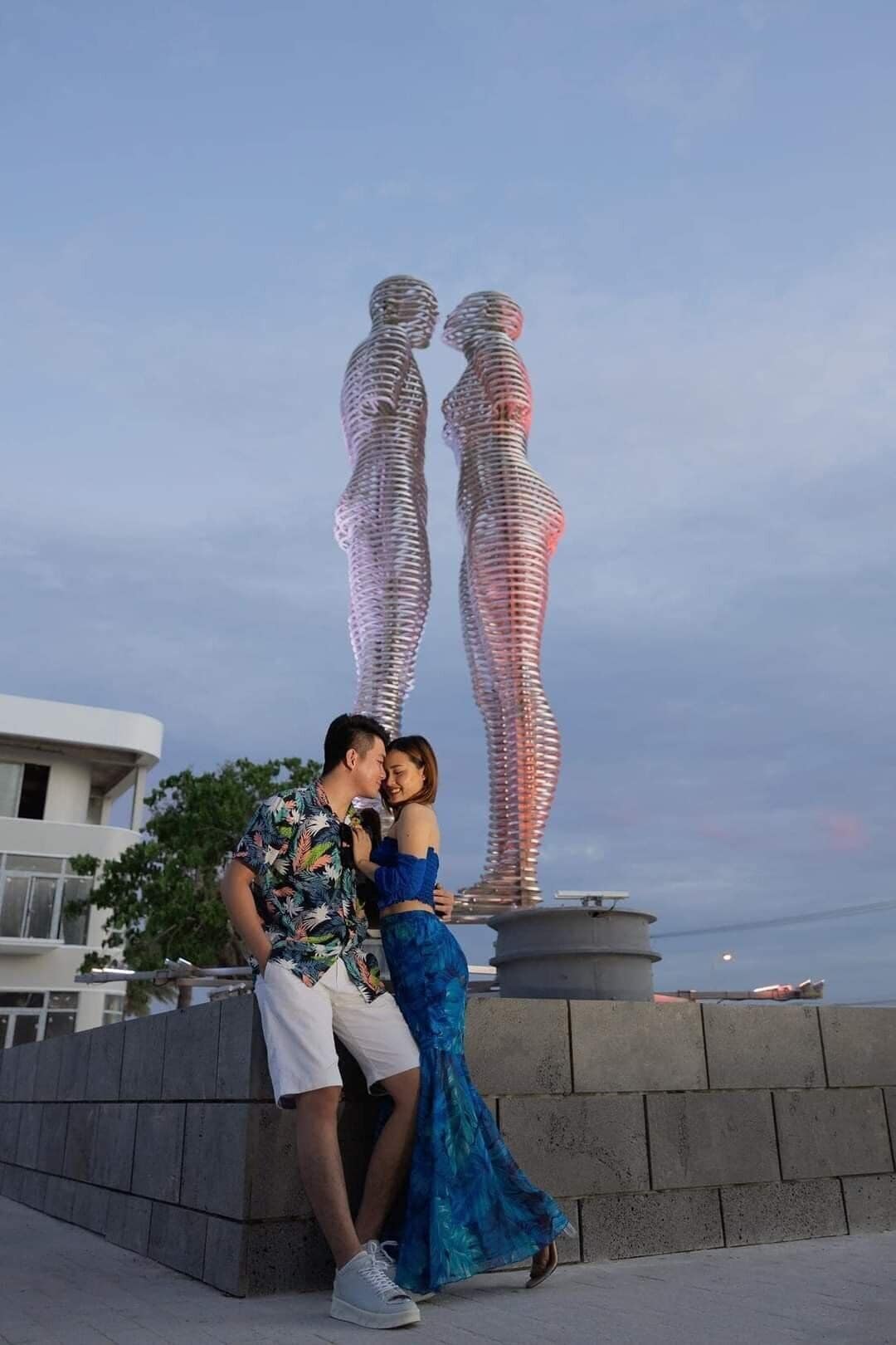 A couple posing in front of a sculpture of two figures.