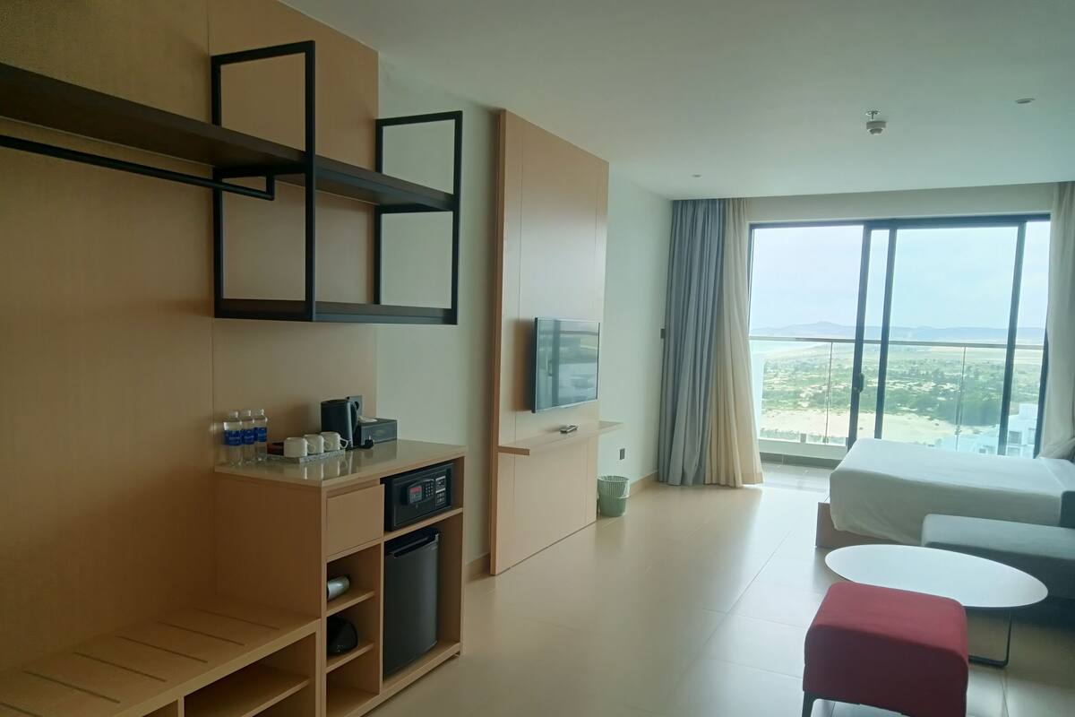 Ett modernt hotellrum med ett stort fönster, en platt-TV, ett skrivbord i trä, en röd pall och en soffa.