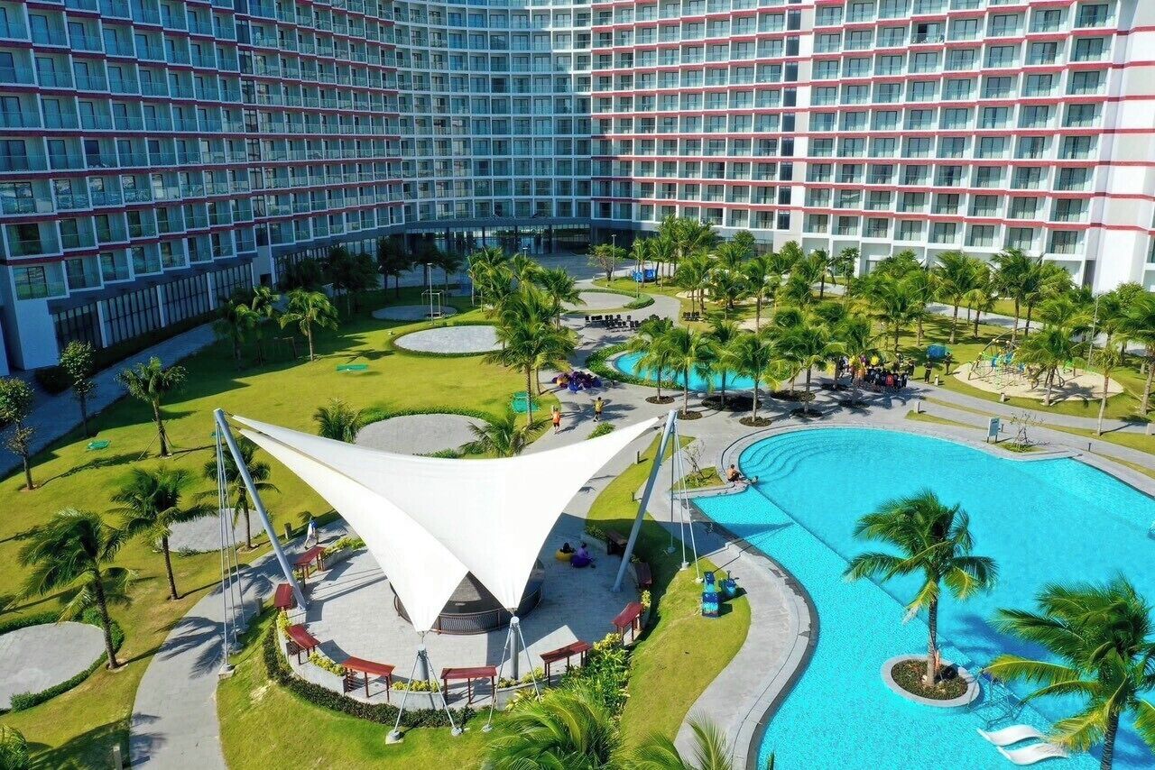Un hôtel doté d’une grande piscine, de palmiers et d’une structure en forme de tonnelle, de couleur blanche.