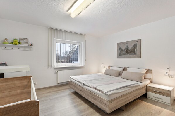 Ein modernes Schlafzimmer mit einem hölzernen Bett, Nachttischen, einem Schreibtisch und einem Fenster mit Jalousien.
