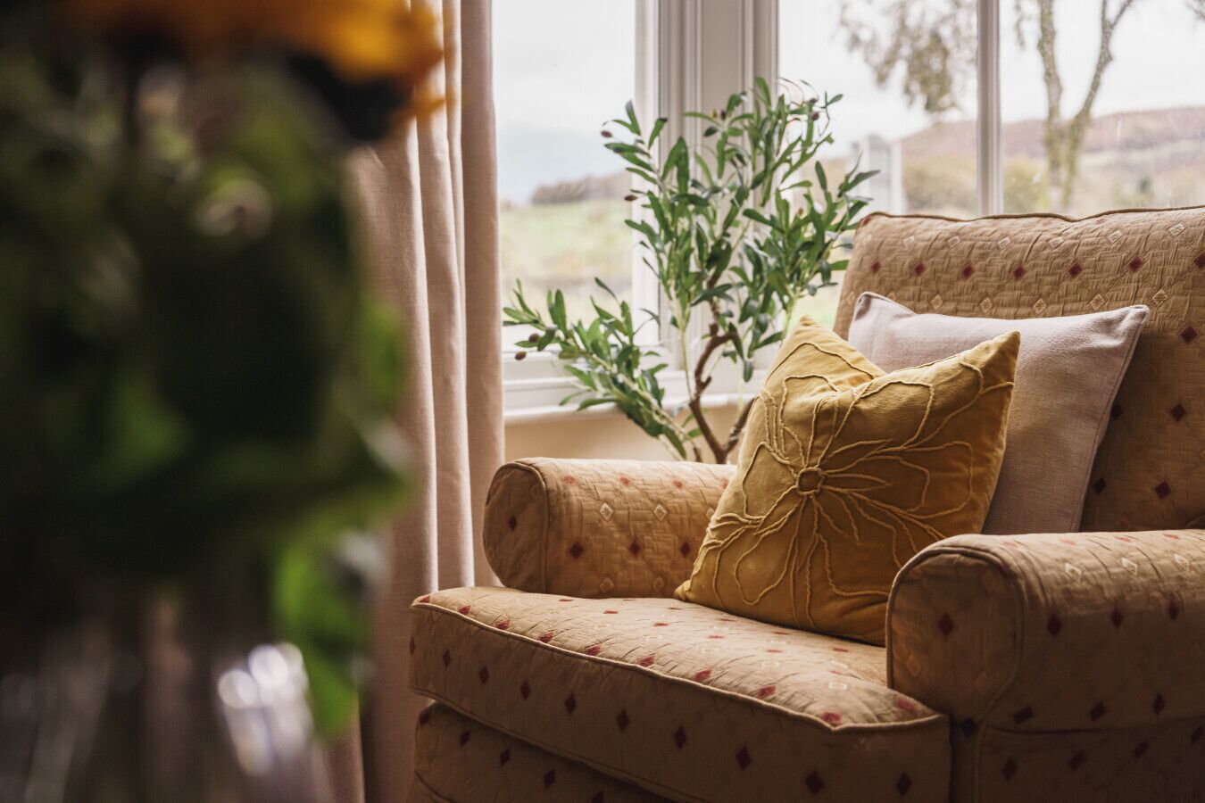 Un sillón cómodo con cojines estampados, un jarrón con flores y vistas al verdor por una ventana.