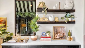 Una sala de estar moderna con una mesa consola blanca, un ventanal con toldo a rayas y estanterías con objetos decorativos y libros.