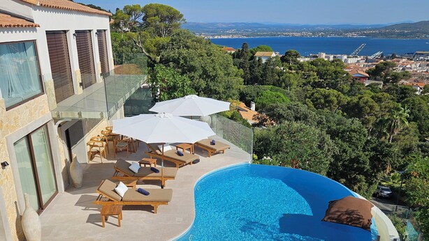 A poolside area with lounge chairs and umbrellas, overlooking a scenic landscape with a view of the sea and distant hills.