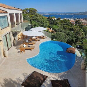 A poolside area with lounge chairs and umbrellas, overlooking a scenic landscape with a view of the sea and distant hills.