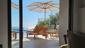 A room with a view of a pool and a beach. There is a wooden lounge chair and a table with an umbrella.