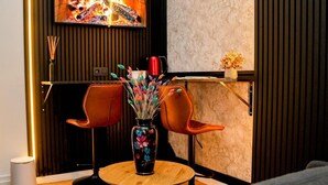 Een gezellige zithoek met twee oranje stoelen, een houten tafel en een vaas met kleurrijke bloemen. Achtergrond: een open haard met vuur.