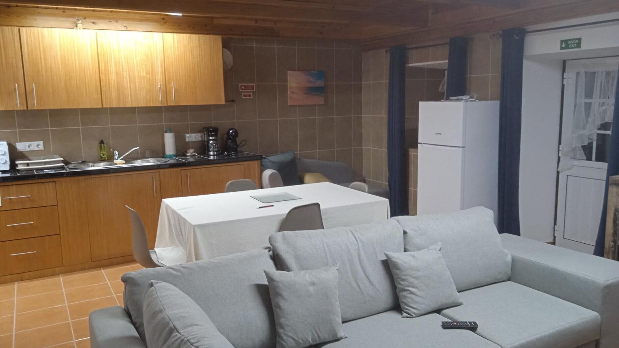 A kitchenette with wooden cabinets, a dining table with a white tablecloth, and a grey sofa with cushions.