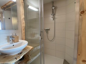 Een badkamer met een douche in een glazen cabine, een witte wastafel op een houten plank en een spiegel boven de wastafel.