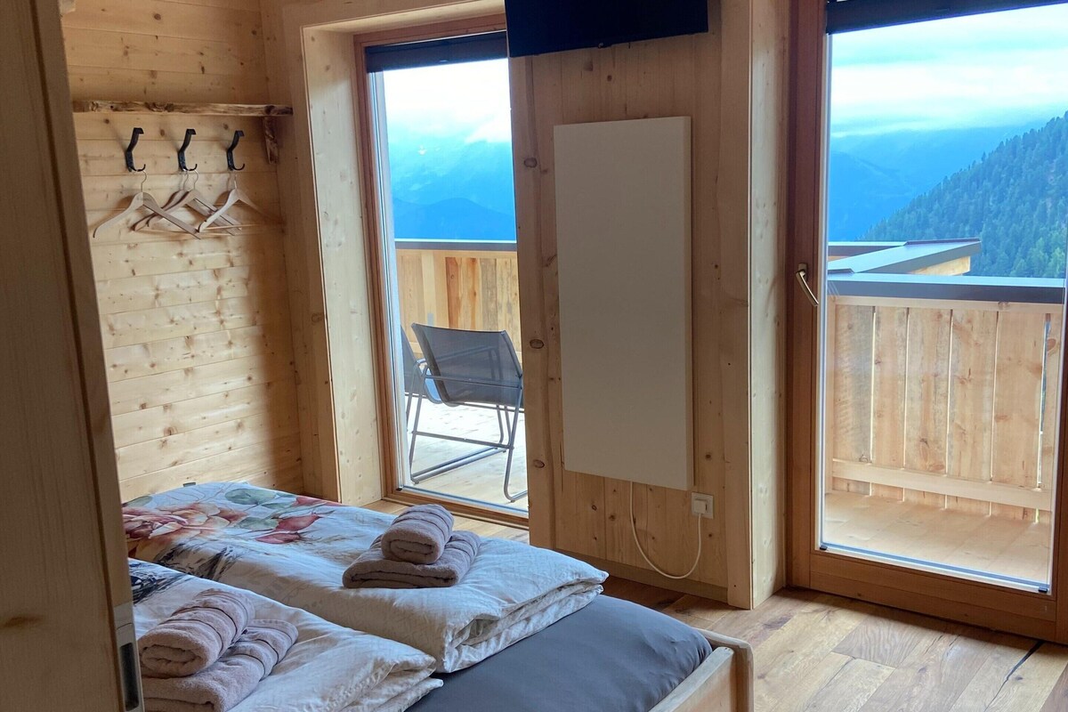 Quarto de cabana de madeira com duas camas, uma cadeira e uma janela grande com vista para as montanhas.