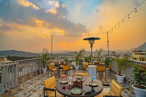 Un área de comedor al aire libre con mesas y sillas, plantas en macetas y luces colgantes sobre un fondo de atardecer.