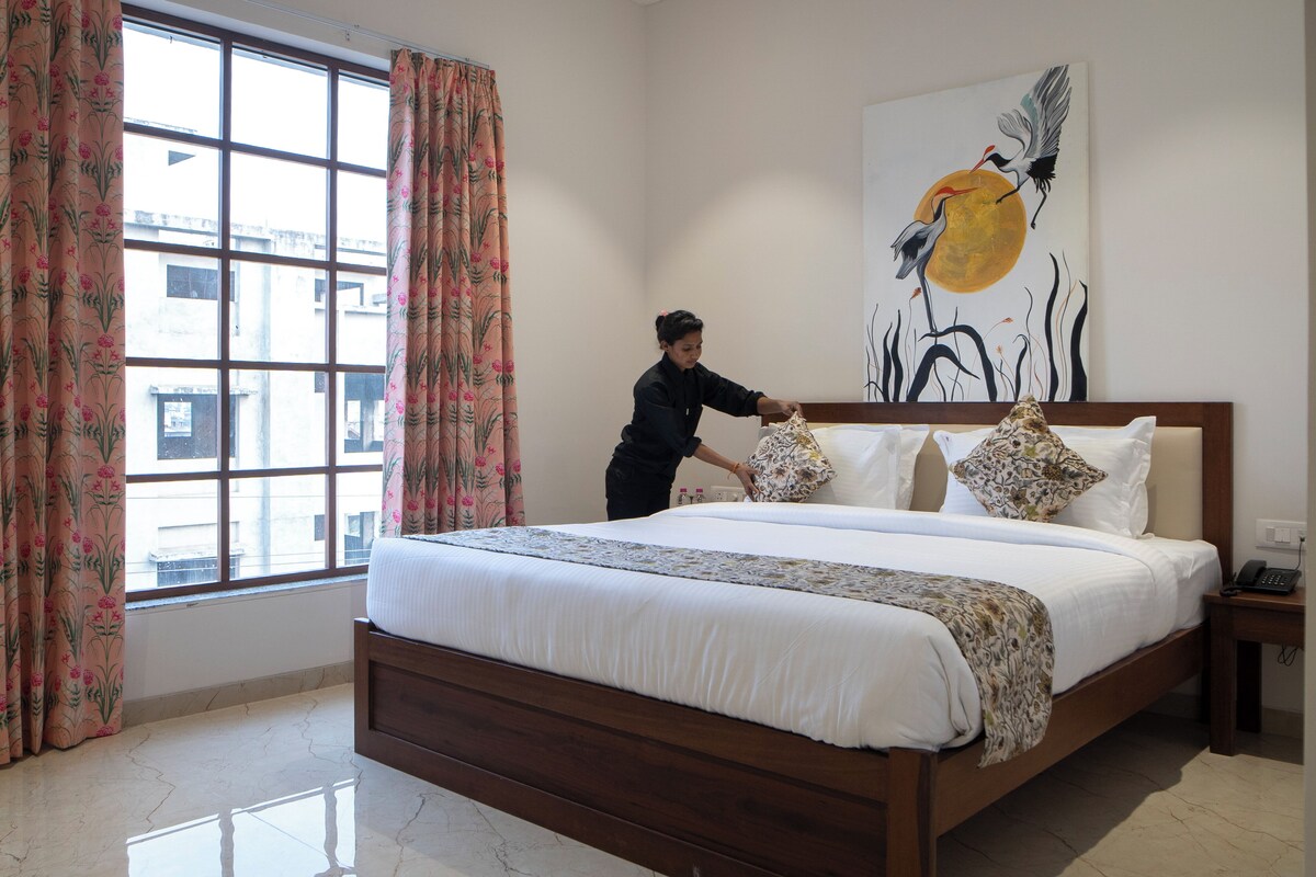 A neatly made bed with floral-patterned pillows, a large window with curtains, and a painting of cranes on the wall.