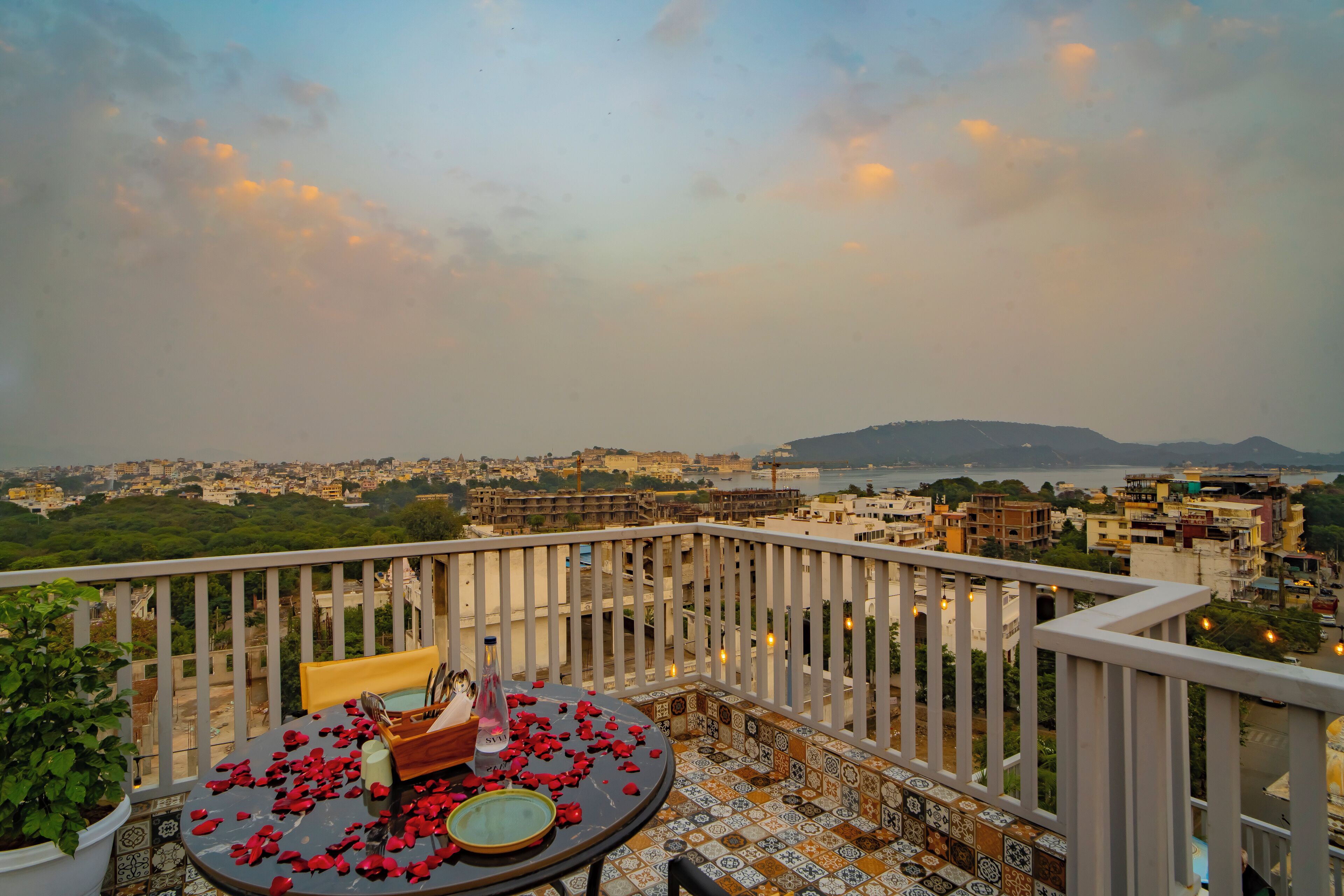 A balcony with a table set for two, overlooking a cityscape with buildings and a distant mountain.