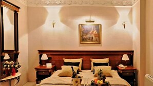 Habitación de hotel con una cama grande, dos mesitas de noche con lámparas, un cuadro en la pared y una cesta de frutas sobre la cama.