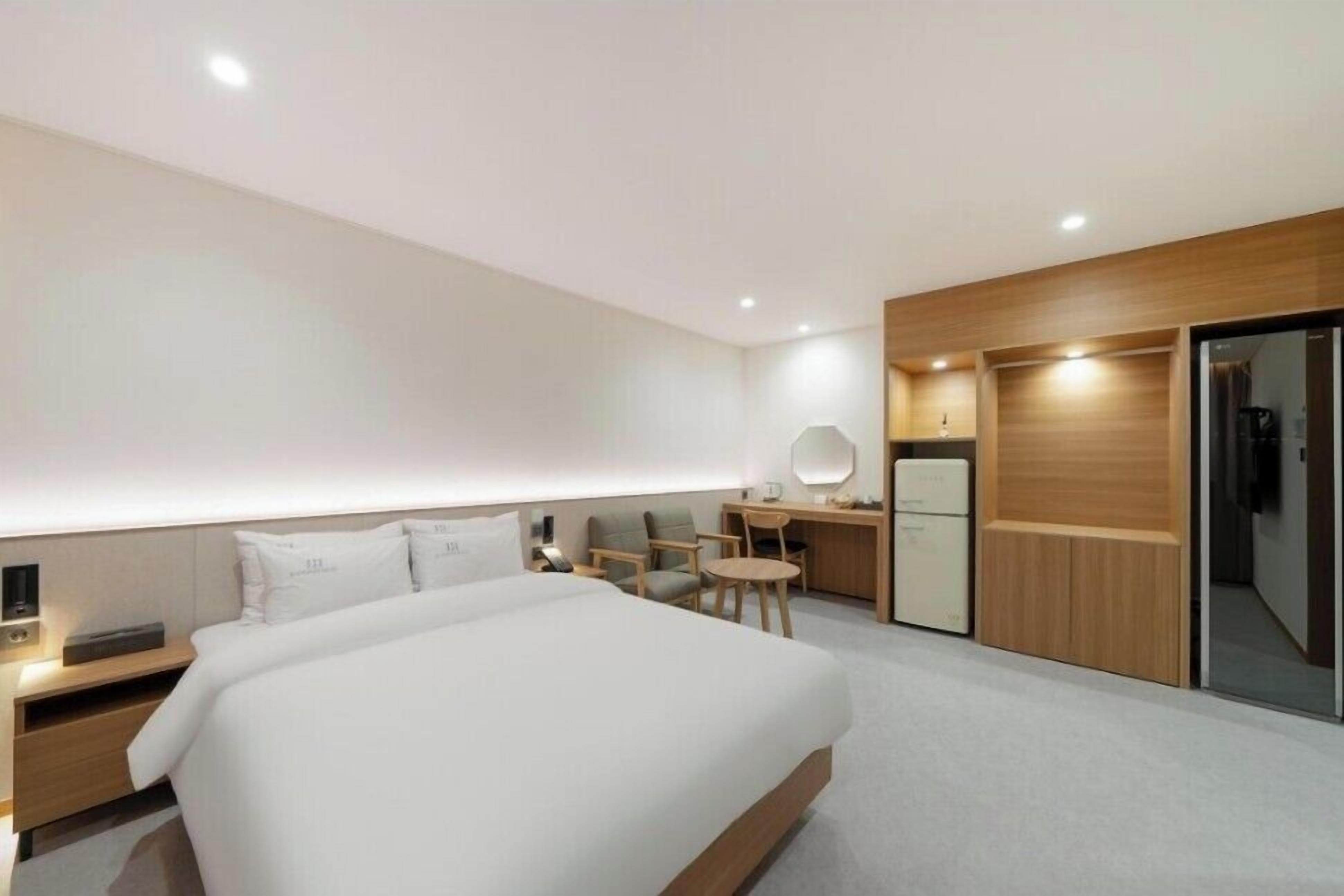 A modern hotel room with a large bed, a desk with a chair, a small round table, and a wardrobe.