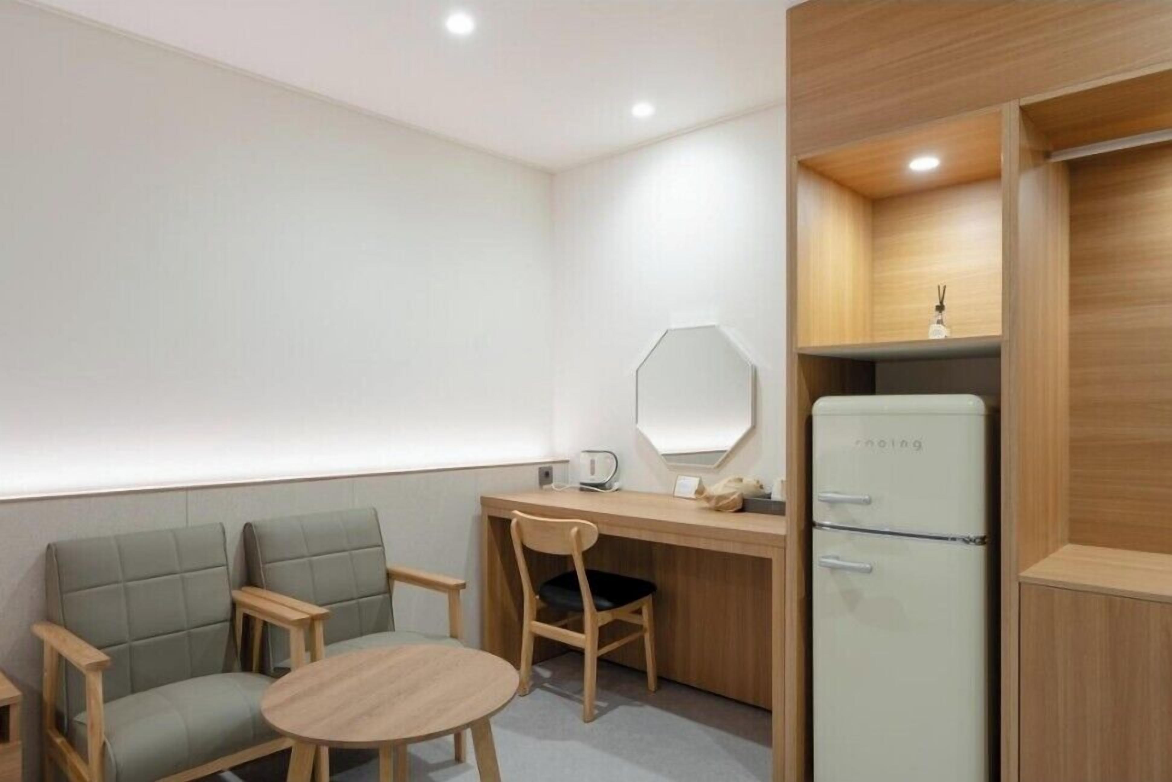A compact room with a wooden desk, a chair, a small round table, a mirror, a refrigerator, and a light fixture.