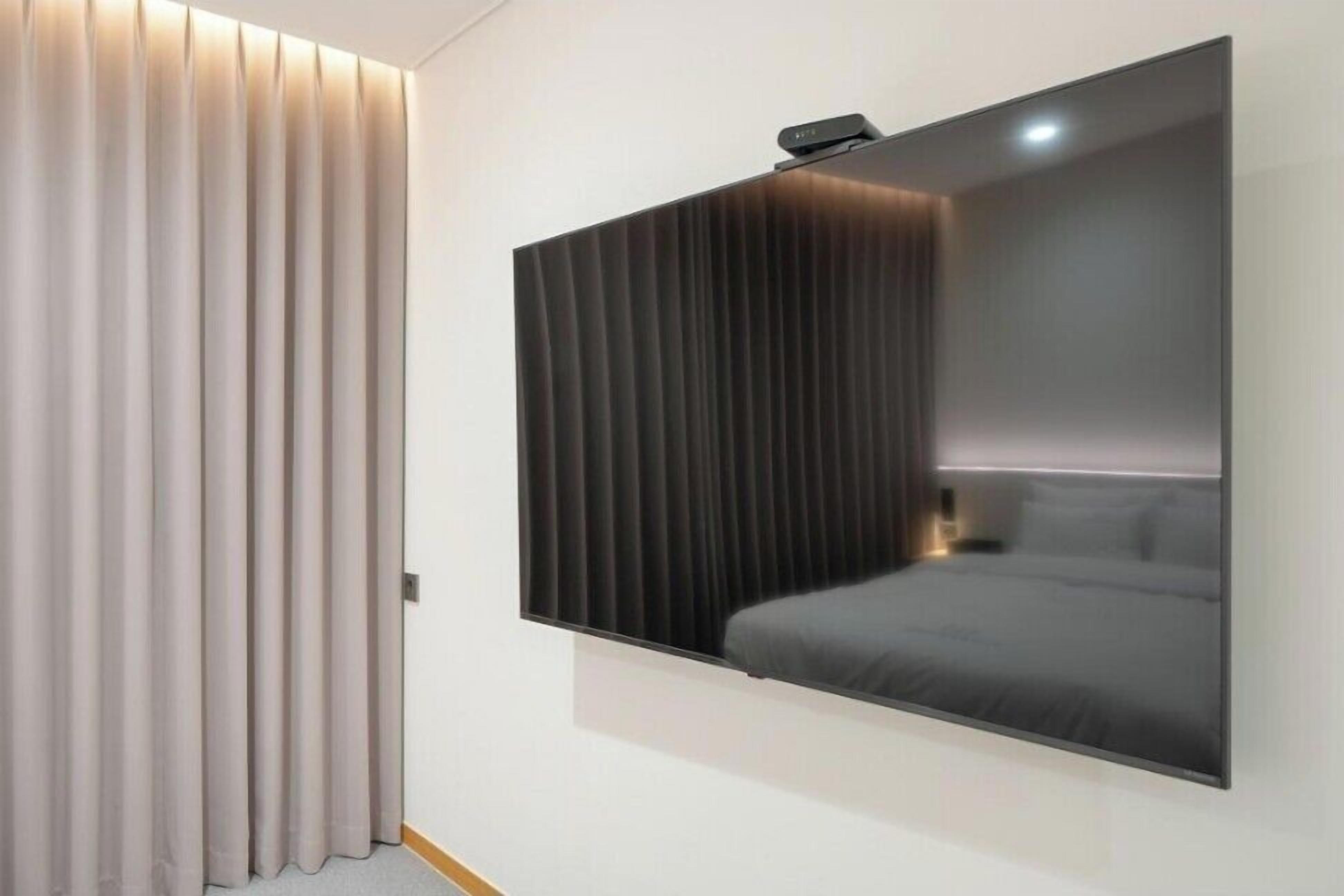 A modern hotel room with a large wall-mounted TV, a bed with grey bedding, and a curtain on the left.