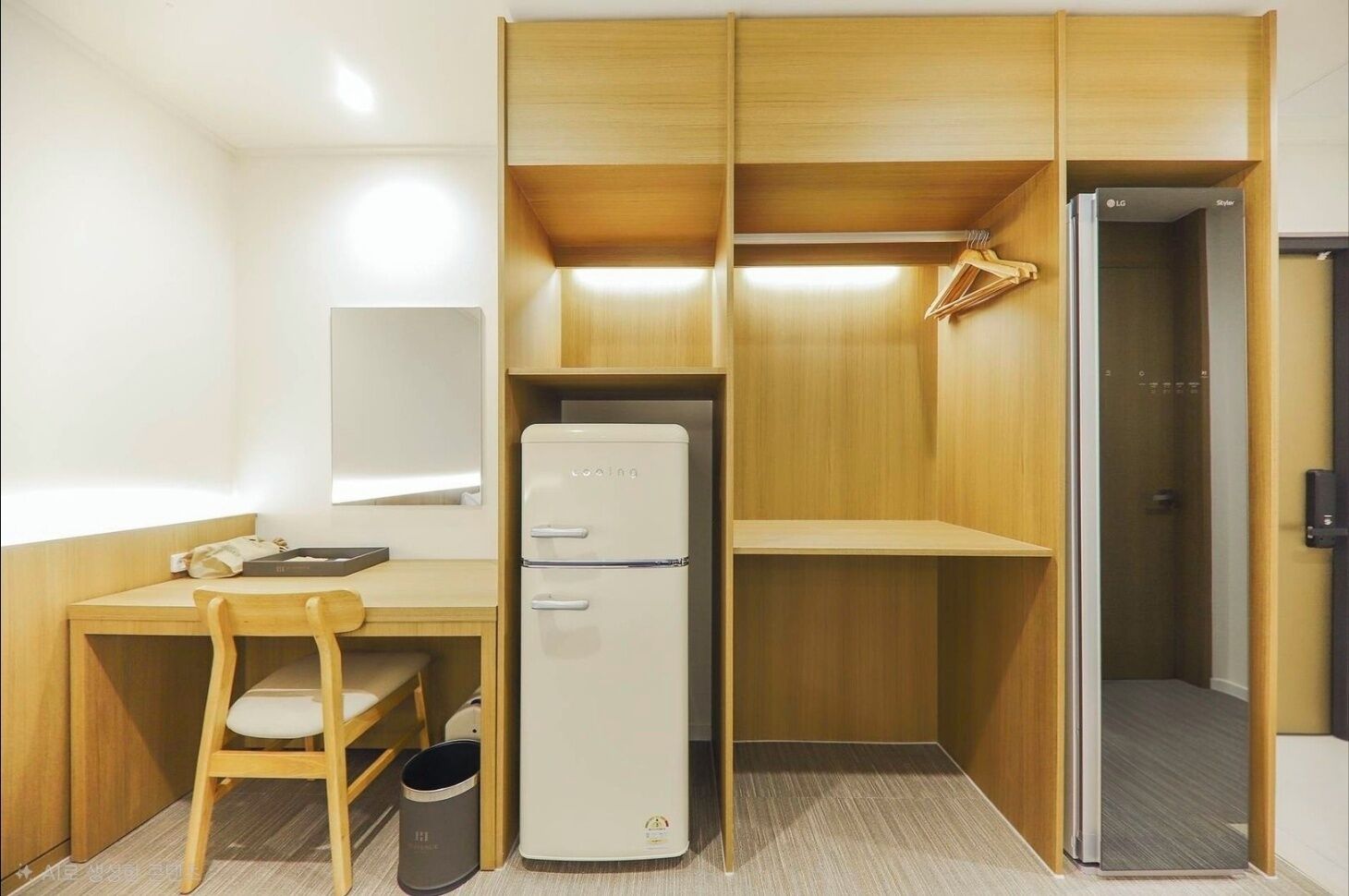 A compact room with a wooden desk, a white refrigerator, a chair, a small trash bin, and a wardrobe with a mirror.