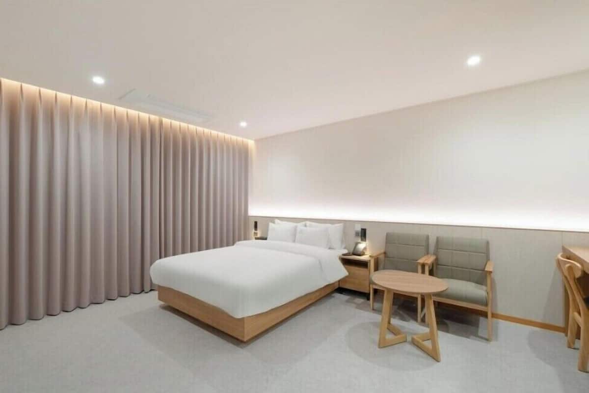 A modern hotel room with a large bed, a seating area with a table, and a large window with curtains.