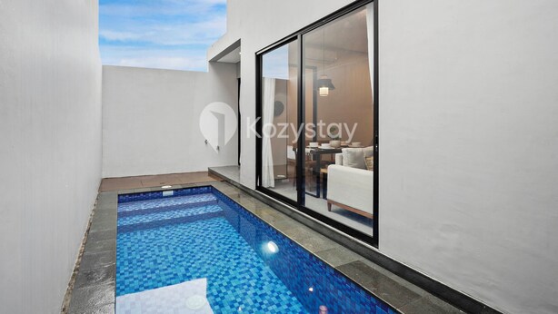 A modern outdoor swimming pool with a tiled floor, adjacent to a white wall and a glass door leading to a living area.
