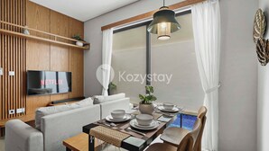 A modern living room with a sofa, dining table set with plates and bowls, a TV mounted on a wooden wall, and a large window with curtains.