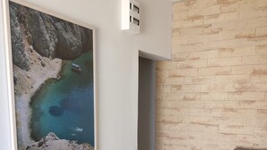 Un pasillo con pared de piedra, un cuadro enmarco de paisaje costero, una silla y una puerta que da a otra habitación.