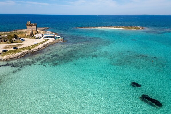 An aerial view of a coastal area with a clear beach, a small tower, and a building with a blue roof.