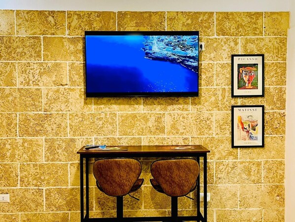 Un televisor montado en una pared con textura, dos cuadros enmarcados y una pequeña mesa con sillas.
