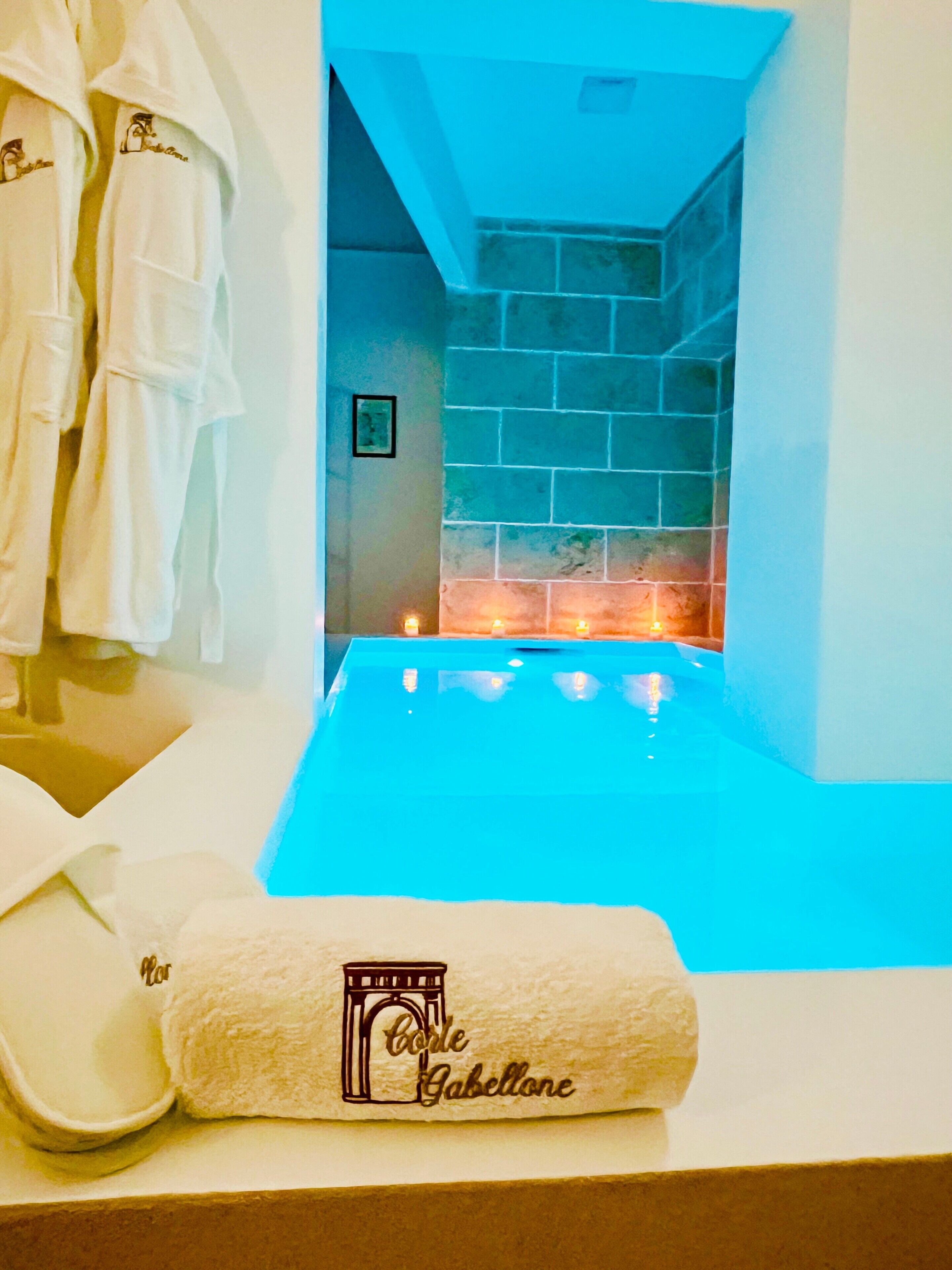 A spa room with a blue heated pool, a towel with 'Corte Gabellone' logo, and robes hanging on the wall.