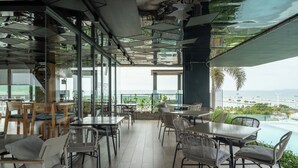 An outdoor dining area with tables and chairs, a view of the sea, and a modern architectural design with glass walls and a ceiling with geometric patterns. - Seaphere residence by Prop Hopper (Sattahip)