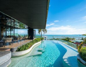 A modern outdoor pool area with a view of the ocean, surrounded by lounge seating and a glass-enclosed bar.