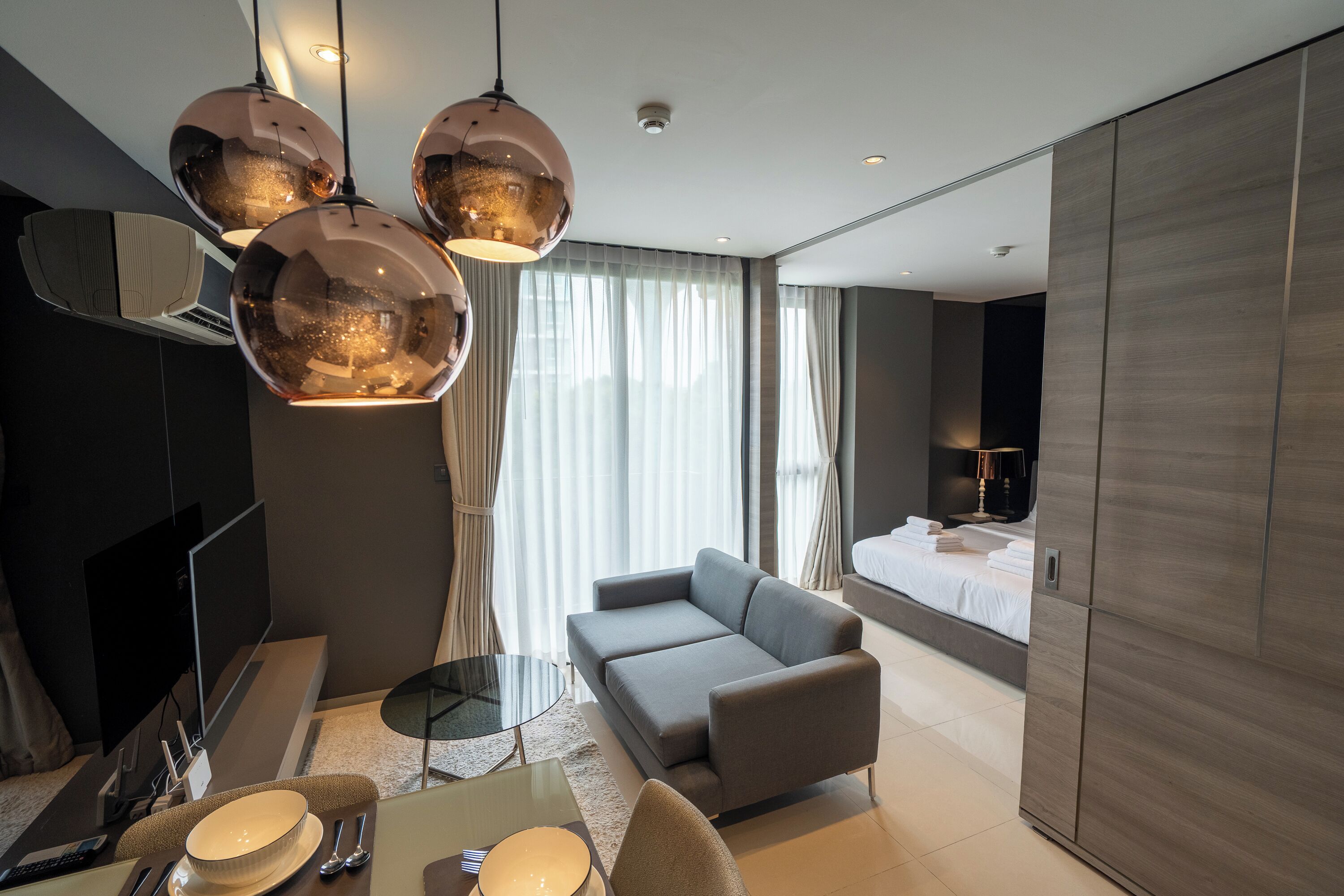 A modern hotel room with a sofa, a dining table with a bowl and cutlery, a TV, and a large bed.
