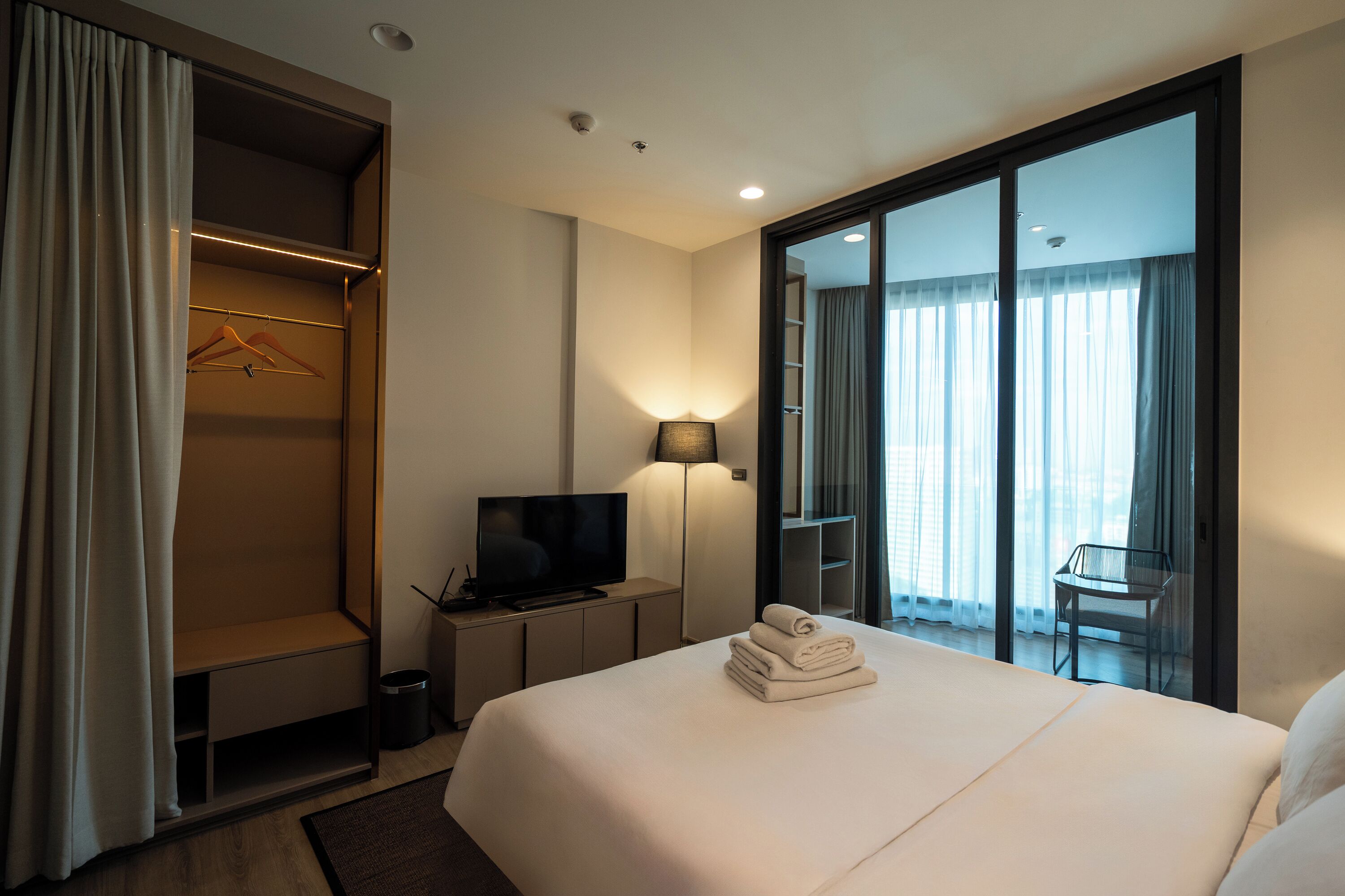 A hotel room with a bed, a TV, a desk with a lamp, a closet, and a sliding glass door to a balcony.