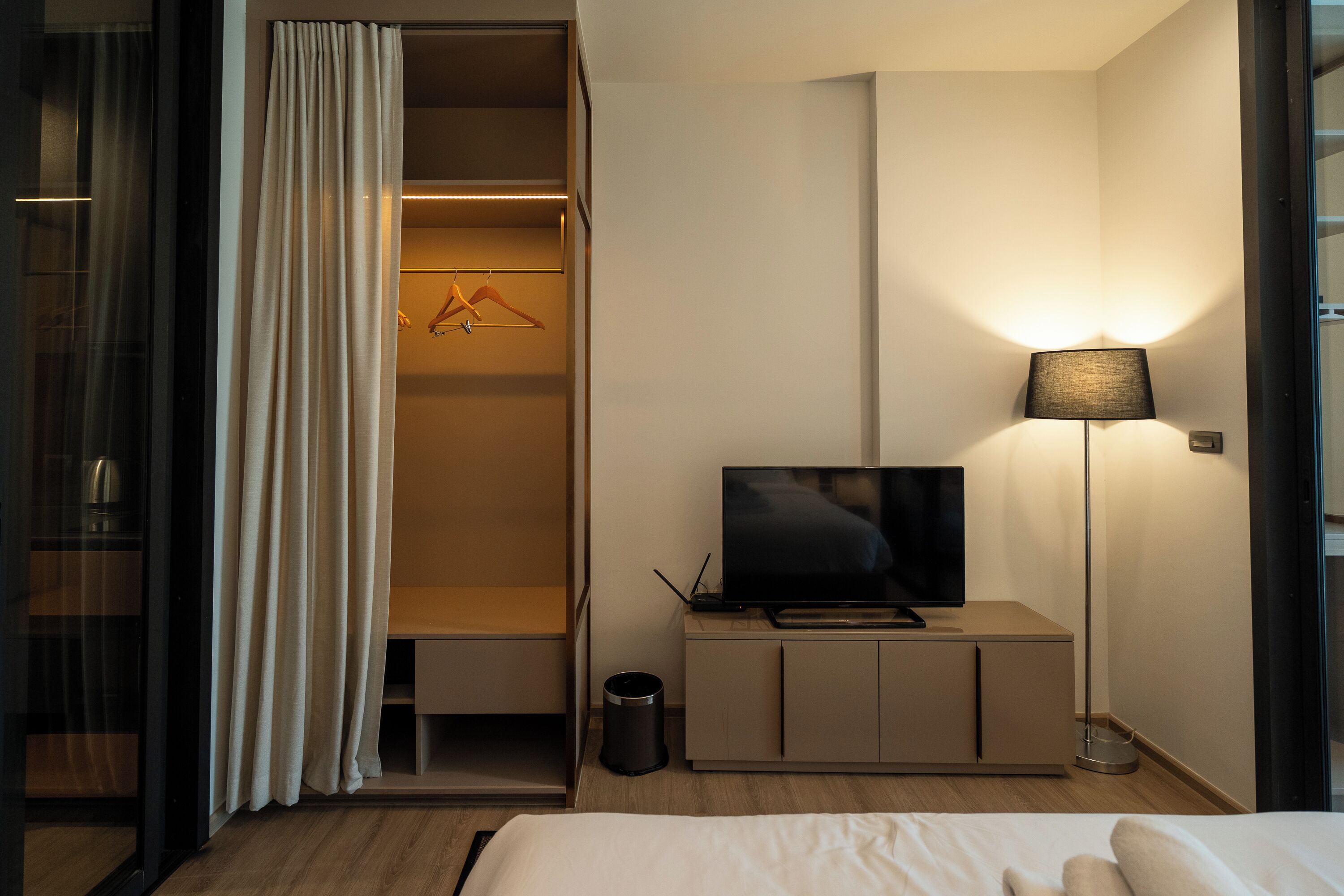 Ein Hotelzimmer mit Bett, Fernseher auf einem Schrank, Kleiderschrank mit Kleidung, Lampe und Mülleimer.